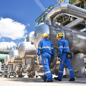 Two engineers in hard hats and blue jumpsuits walking past industrial pipelines and machinery.